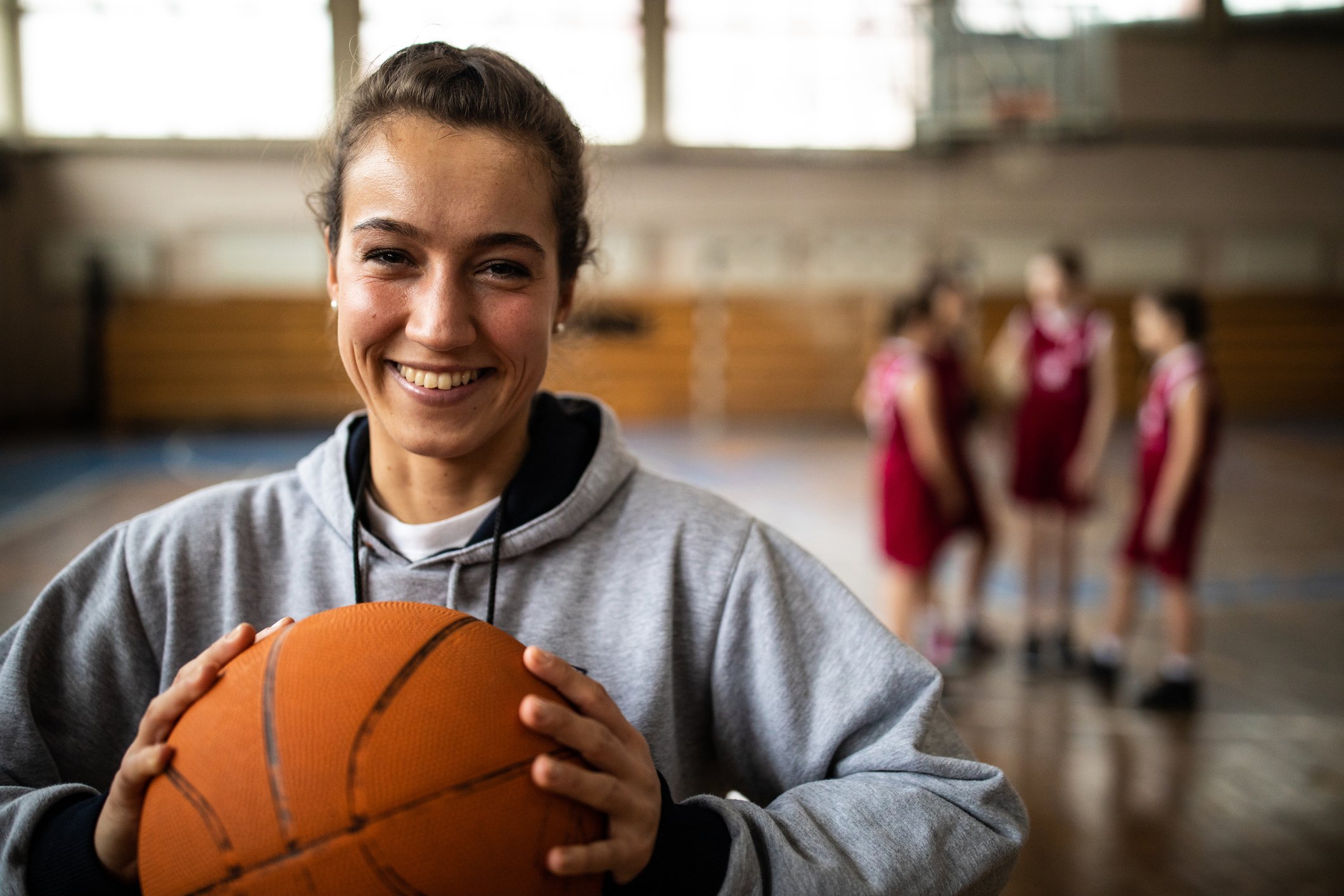 A basketball player on the court holds a ball and smiles as other players stand in the background. 