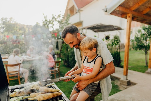 A parent playfully teaches child how to grill in a backyard setting.