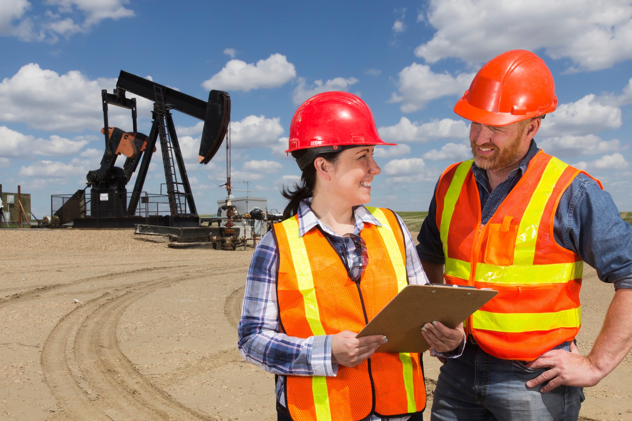 Two oil workers wearing hard hats and vests in front of an oil rig.
