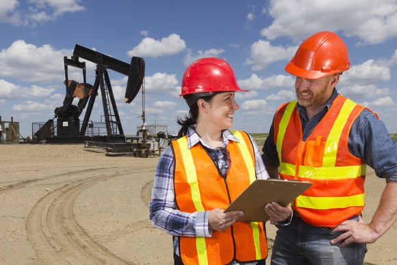 Two oil workers wearing hard hats and vests in front of an oil rig.