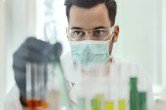 A scientist wearing protective equipment picks up a test tube from a rack while working in a laboratory.