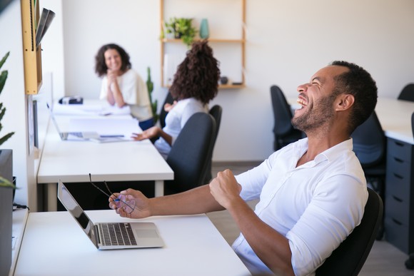 A person leaning back in a chair laughs while sharing an office space with two other people.