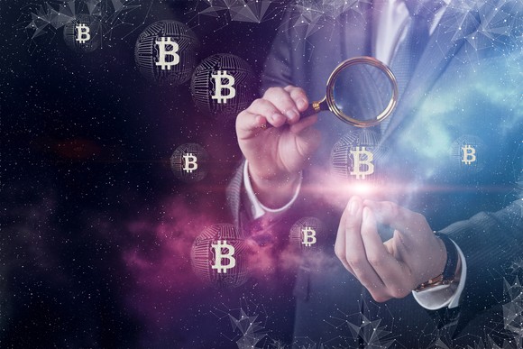Person holding a magnifying glass with Bitcoin tokens floating in the air.