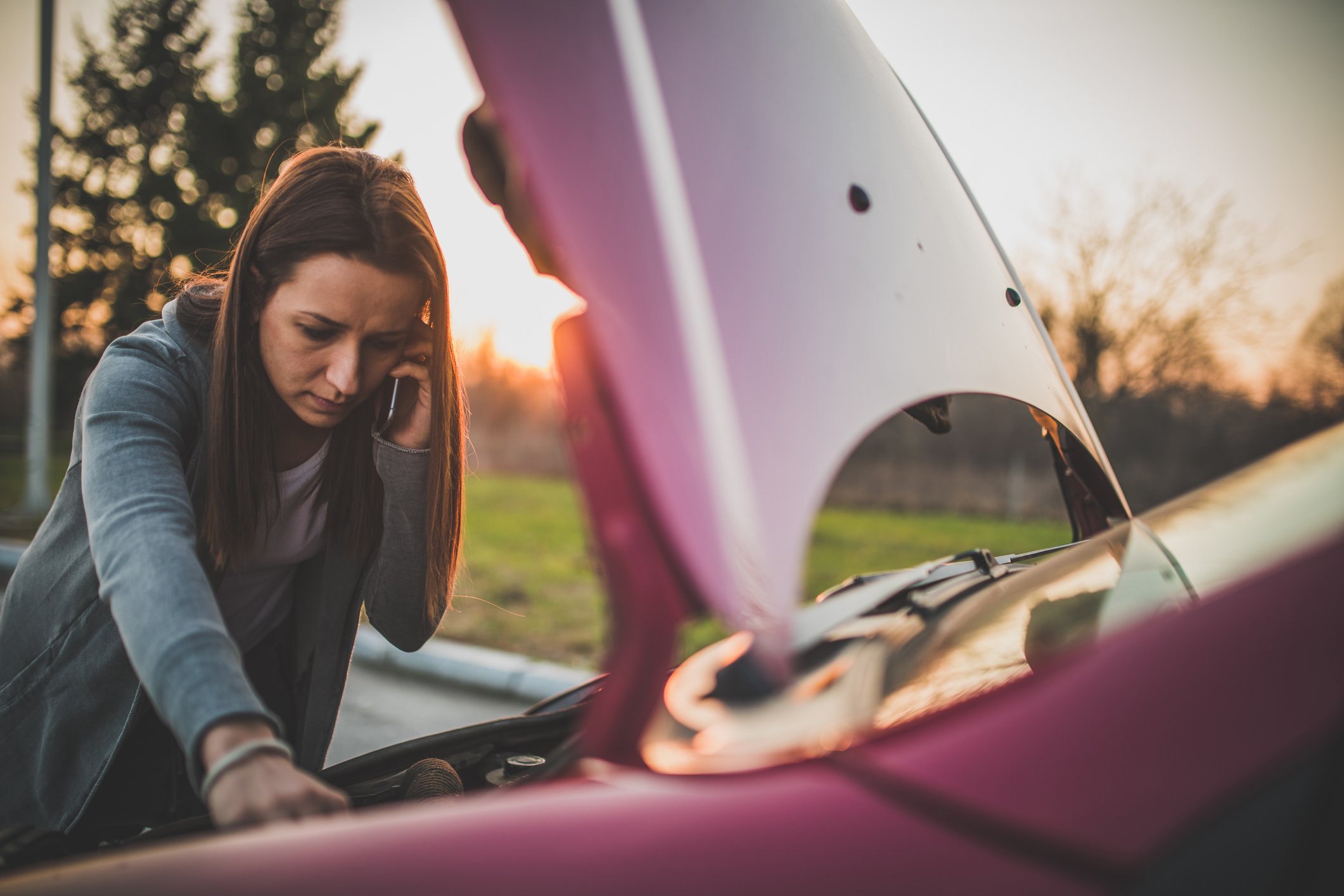 Person looking under open hood of car while using a cell phone.