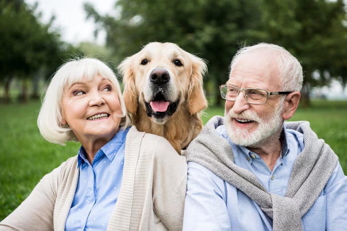 Two older people smile at the park with a dog between them.