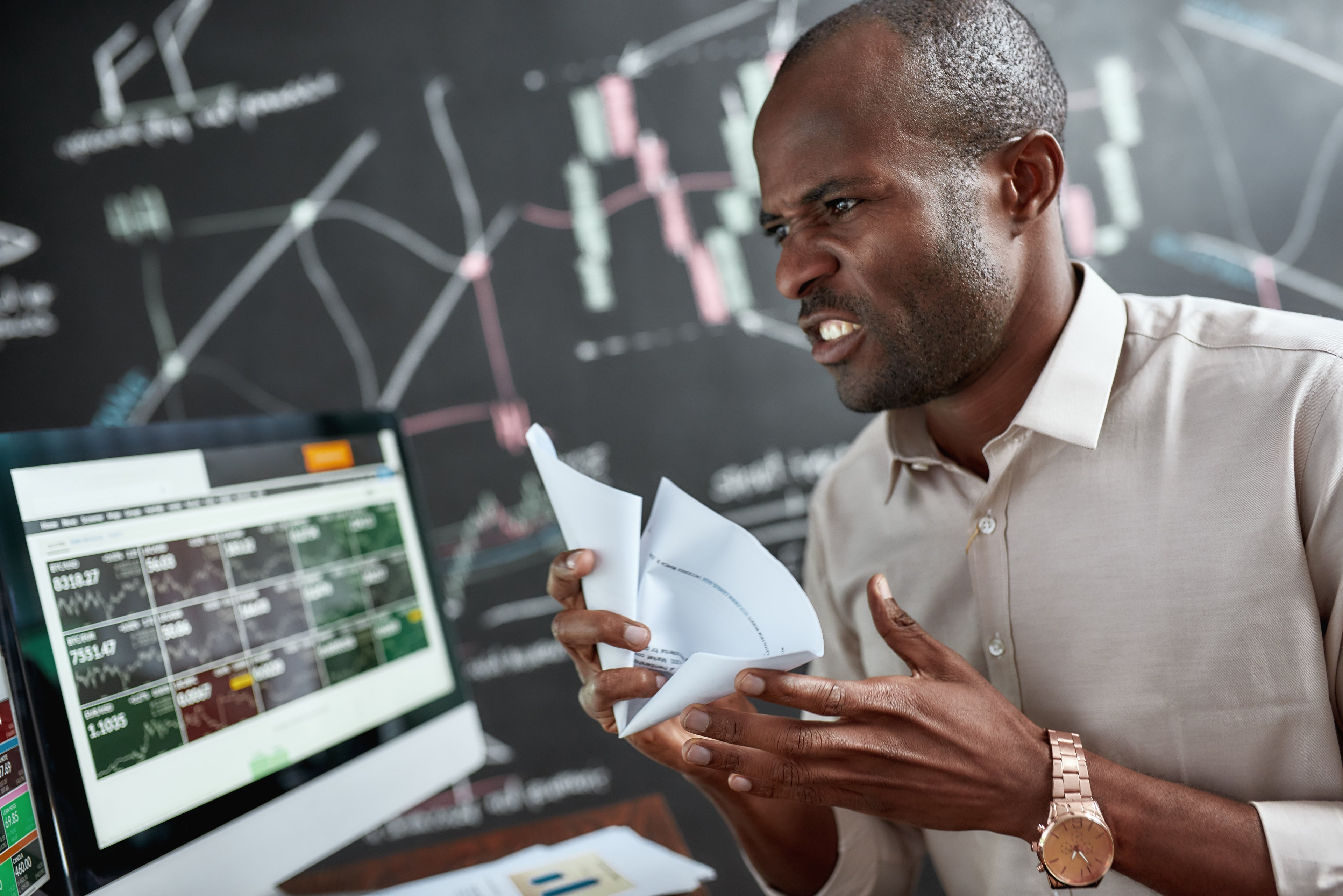An angry trader wearing a cream-colored collared shirt crumples a sheet of paper in anger as stock prices decline in the background. 