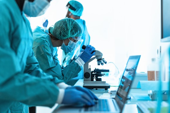 Three medical workers  wearing blue lab gear in a laboratory setting with laptop and microscope.