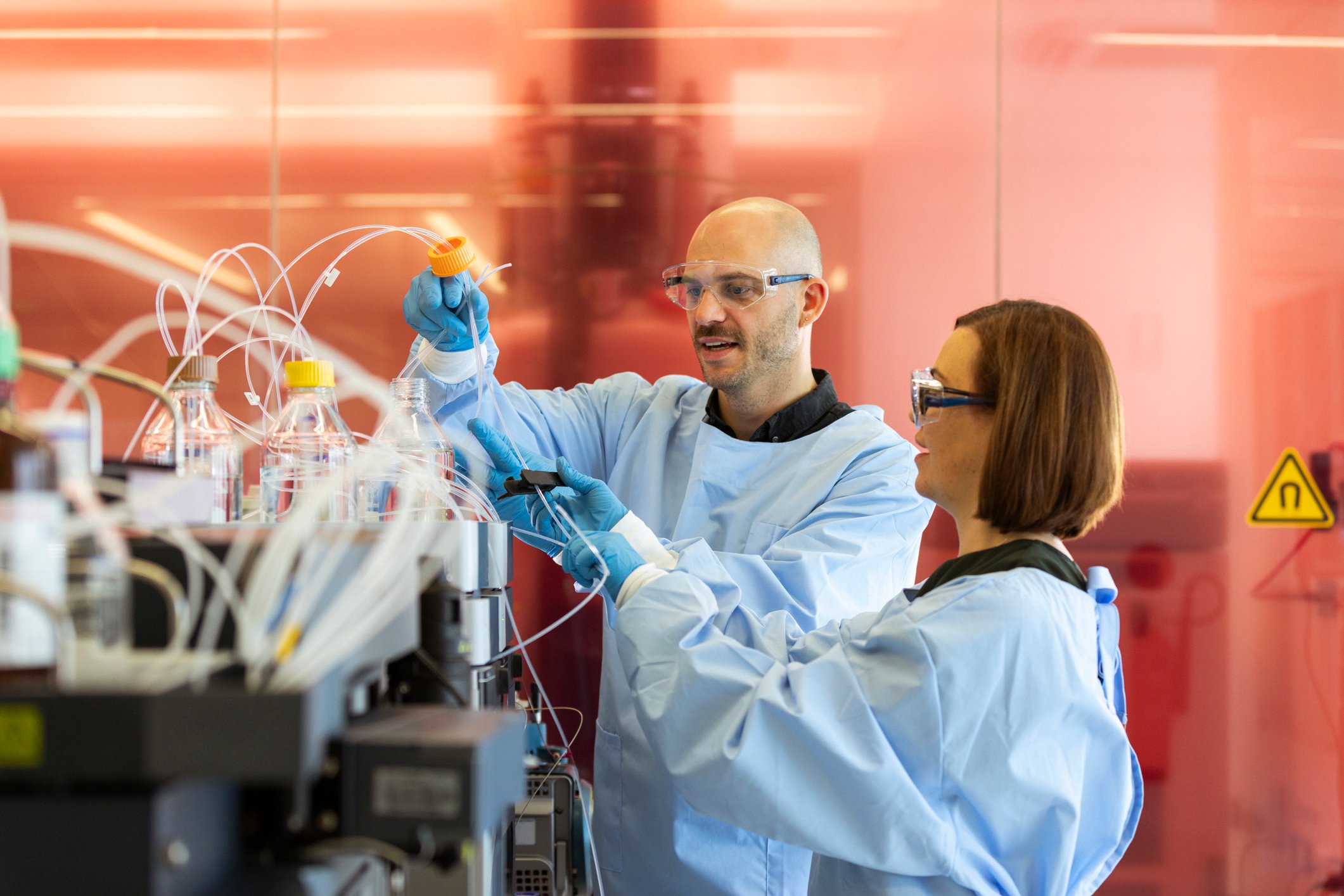 Two scientists chat as one manipulates tubing attached to a complicated biomedical laboratory instrument.