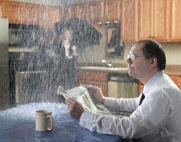 Two people sitting in a kitchen being rained on, one getting wet while reading the paper, the other under an umbrella.