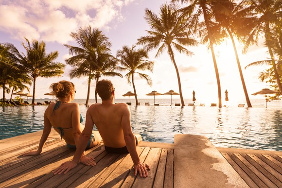 A couple relaxing by a large pool staring at the sun setting over open water and palm trees.