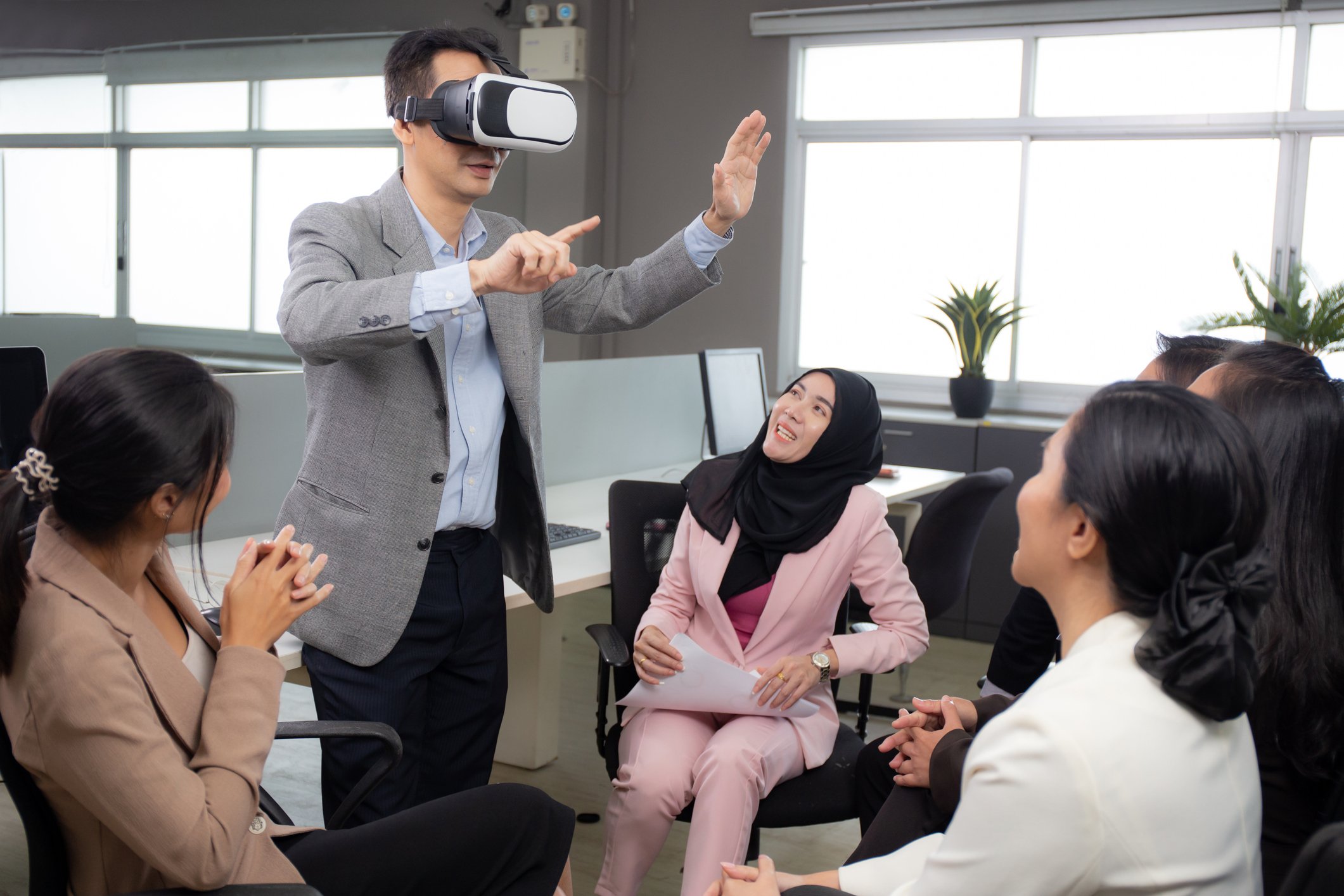 An office worker stands up and wears a VR headset as co-workers observe.