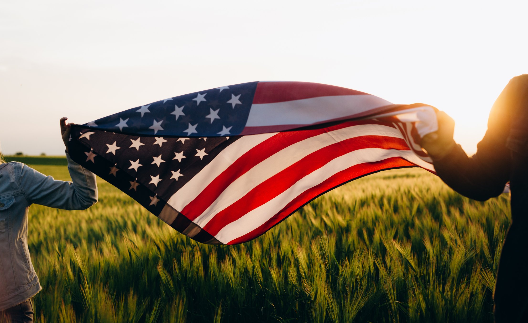 Two people holding an American flag as it waves over a wheat field.