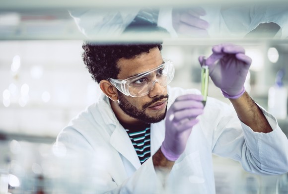 Person examining a tube while wearing gloves, eye protection, and a lab coat.