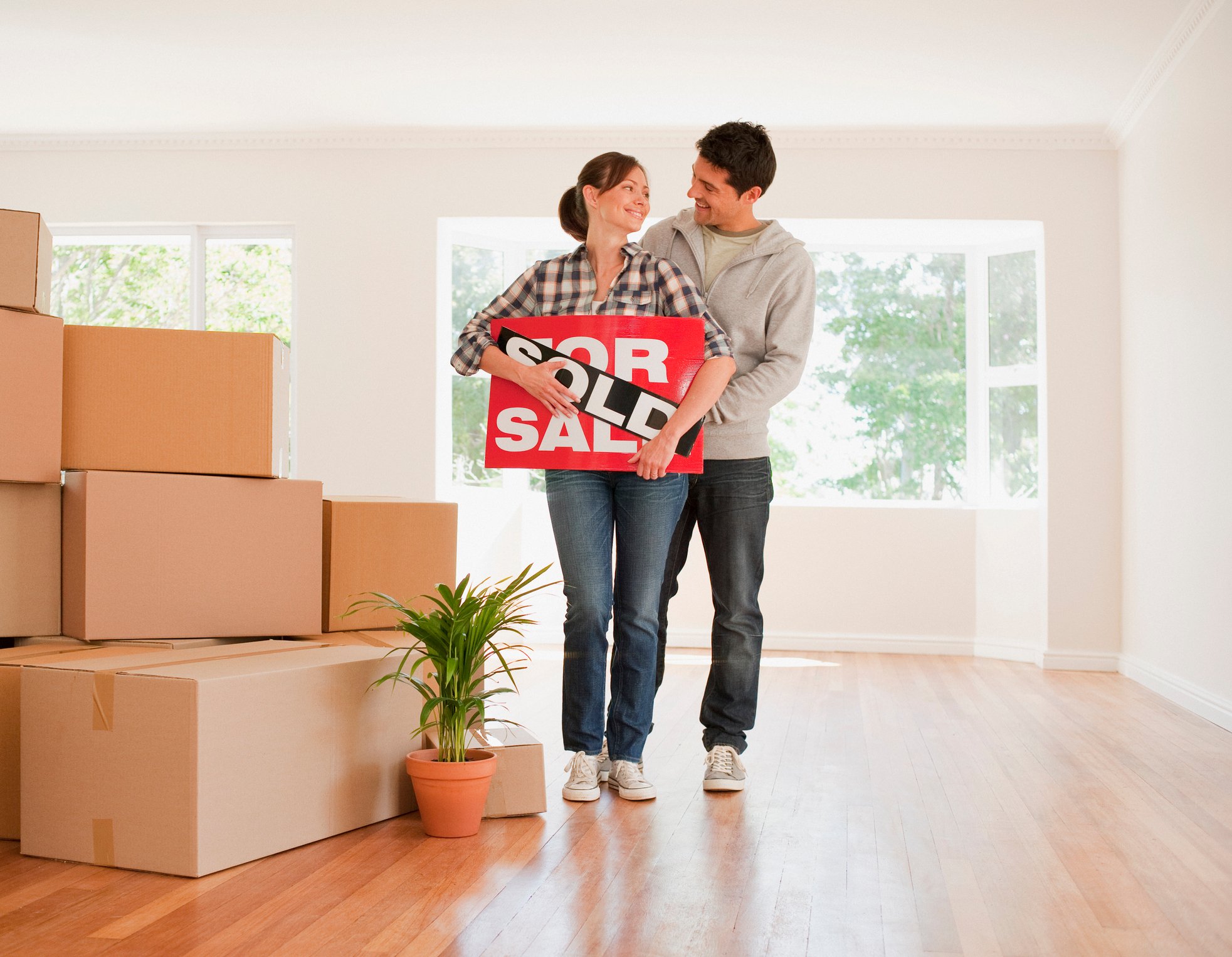 Two people inside a home filled with moving boxes hold a for sale sign with Sold written across it.