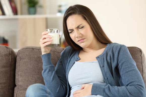 A person frowns and clutches their stomach, holding a cup of milk in their other hand.