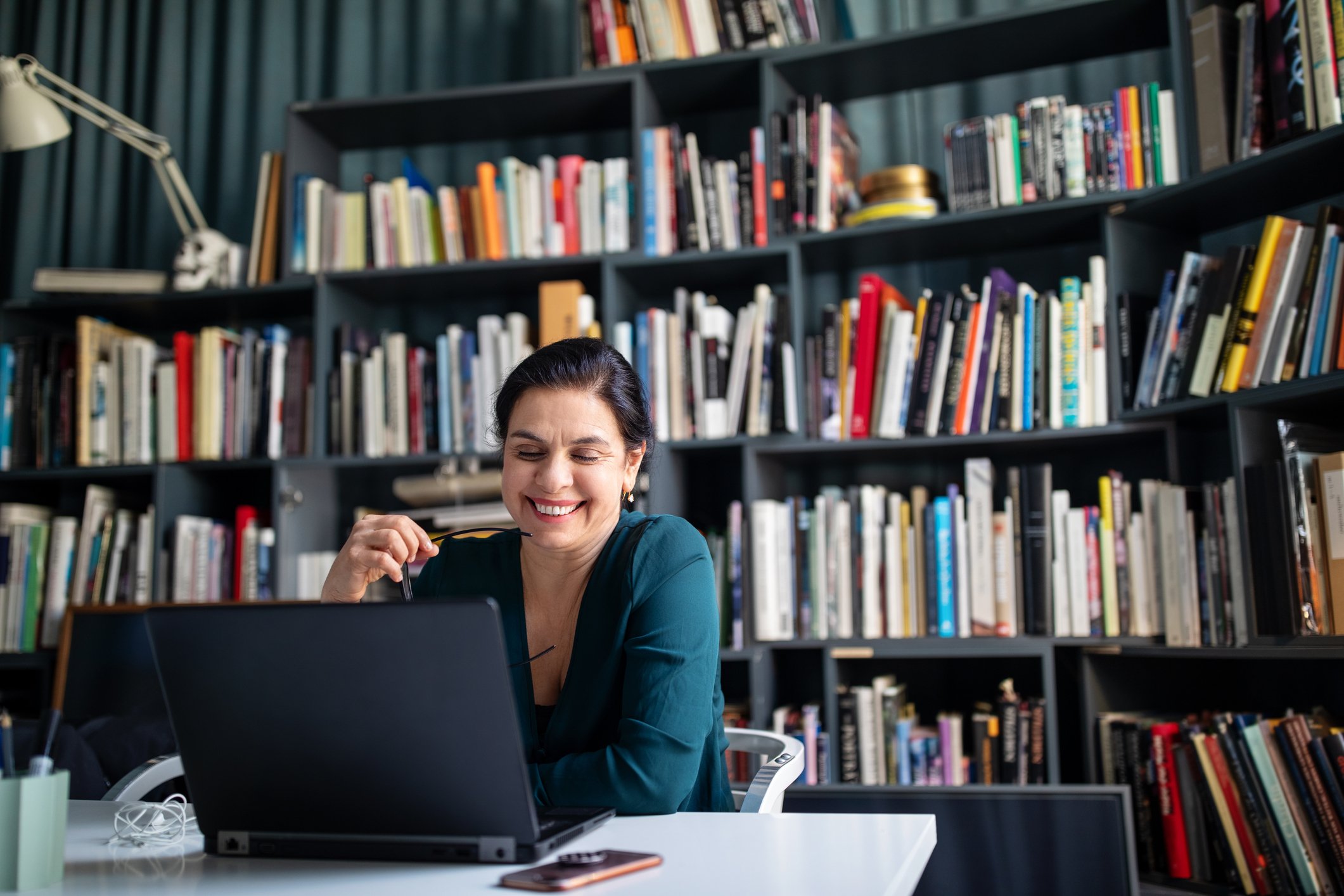 Person looking at a laptop and smiling with bookshelves in the background.