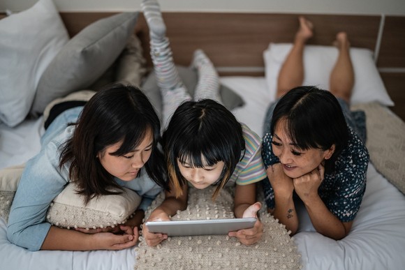 Three people looking at a tablet while lying on a bed.
