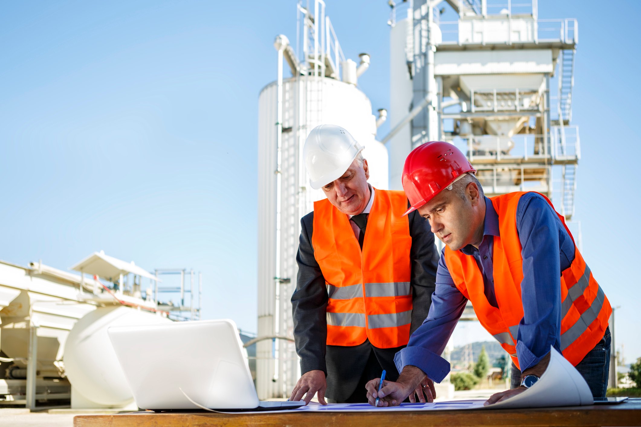 Two men in hard hats and vests looking at blueprints in front of an oil facility.