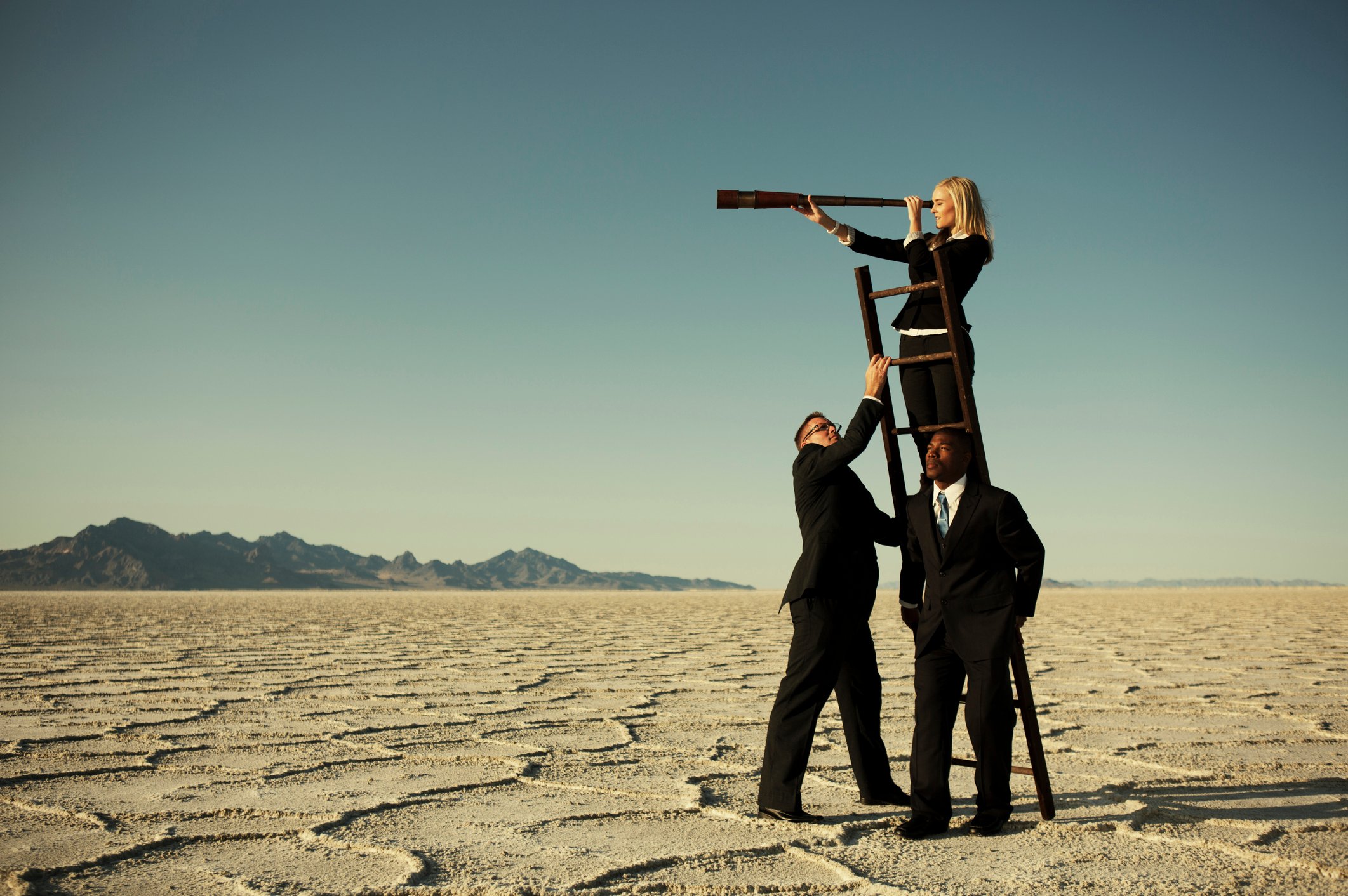 Three people forming a pyramid so the person at the top can look through a telescope a long way off in the distance.
