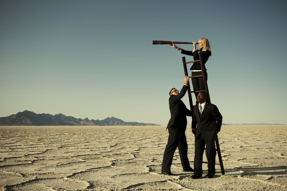Three people forming a pyramid so the person at the top can look through a telescope a long way off in the distance.