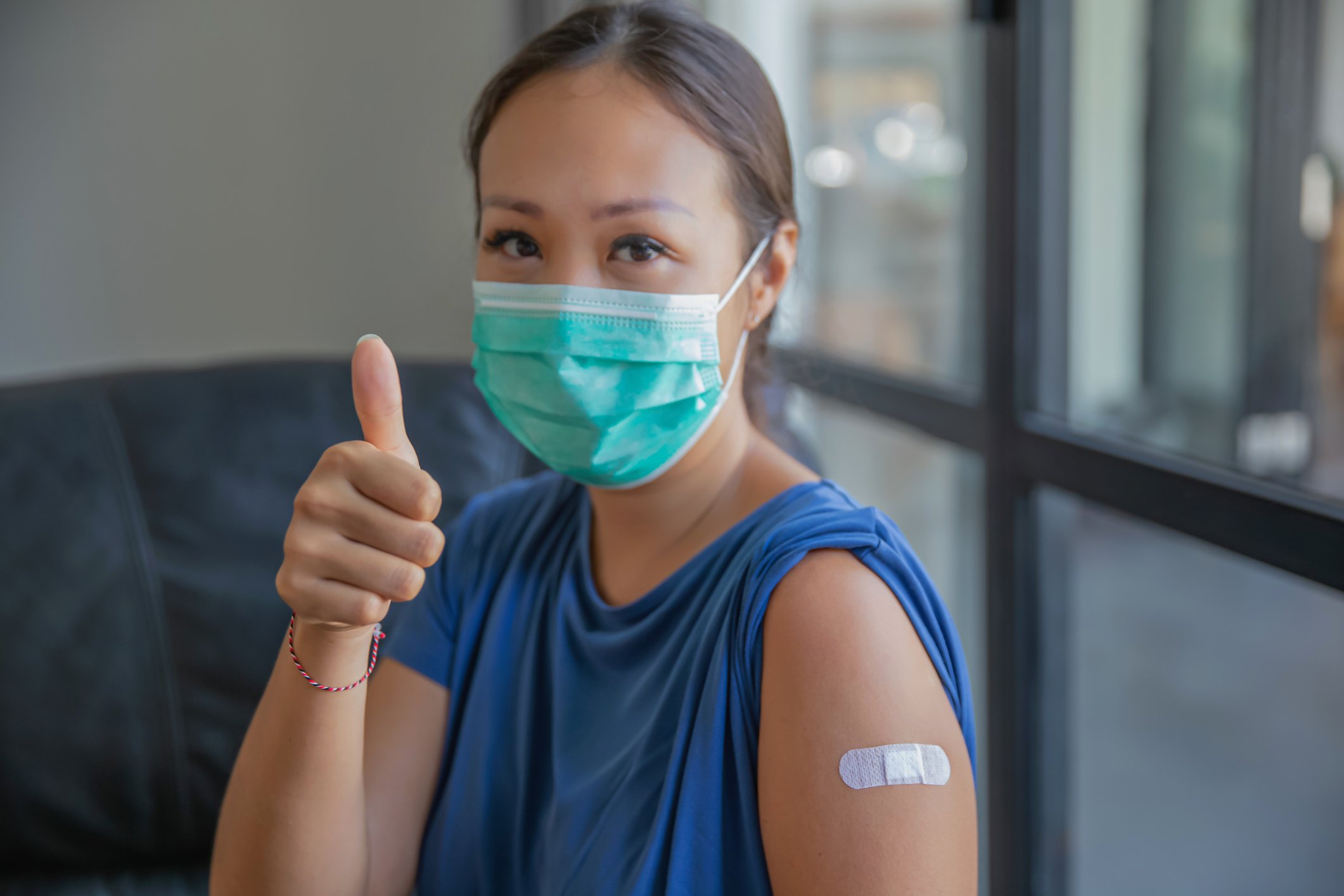 Person in surgical mask with band aid on arm gives the thumbs-up sign.