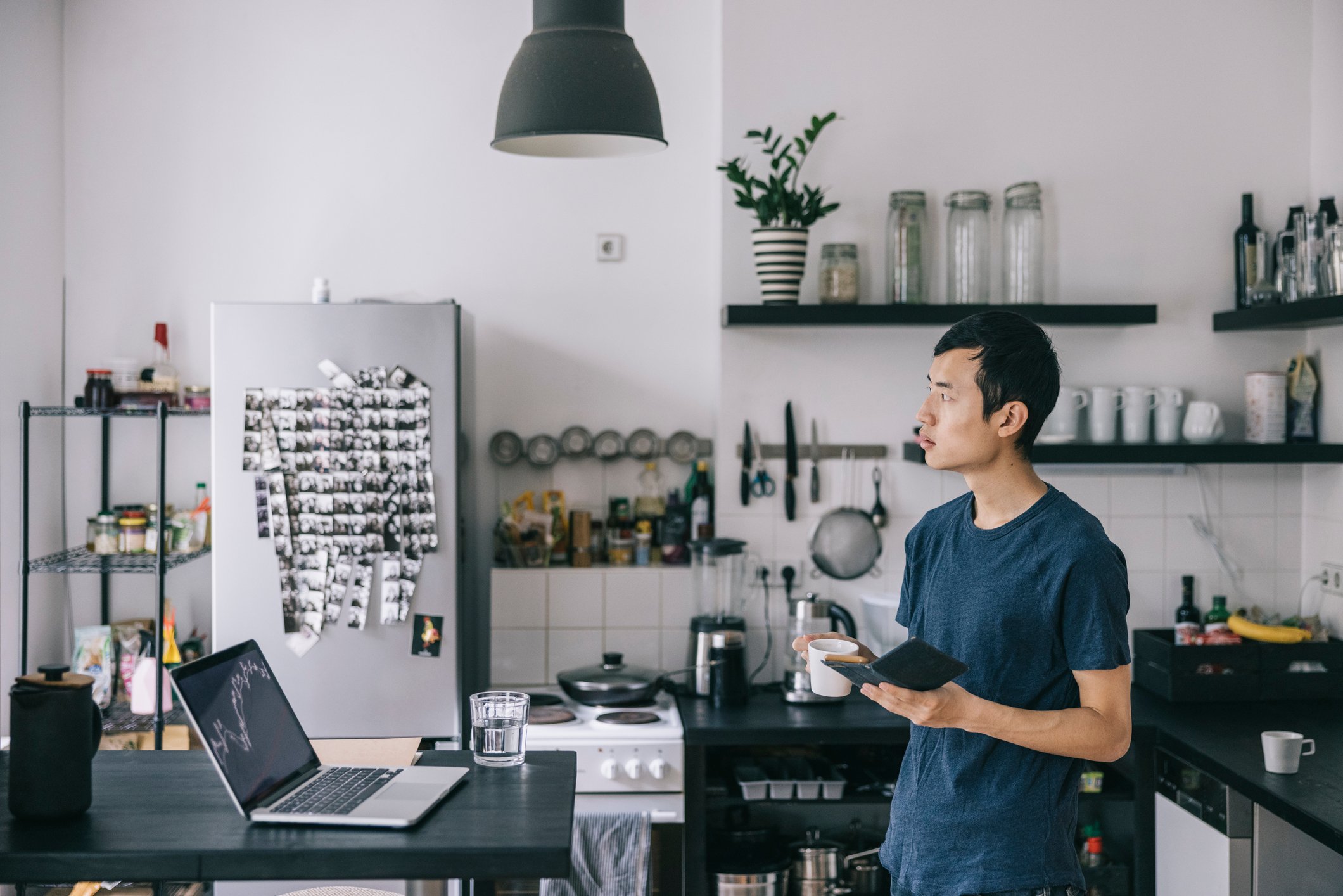 Person looking up as they stand in the kitchen in front of a laptop with a notebook in hand. They also have a coffee.