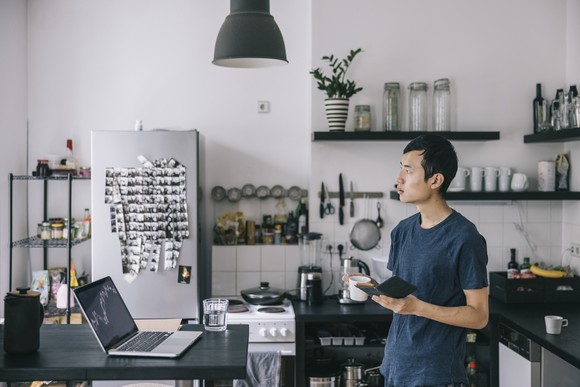 Person looking up as they stand in the kitchen in front of a laptop with a notebook in hand. They also have a coffee.