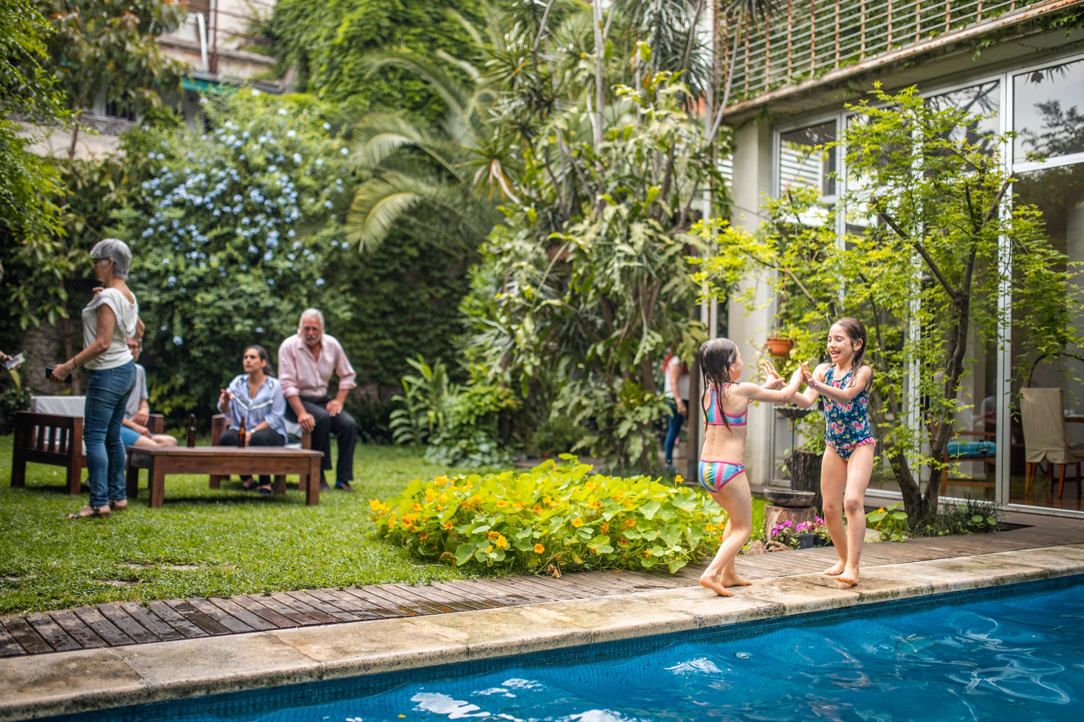 A family and grandparents relaxing in a tropical backyard while young kids play in the pool.