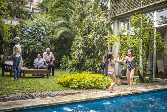 A family and grandparents relaxing in a tropical backyard while young kids play in the pool.