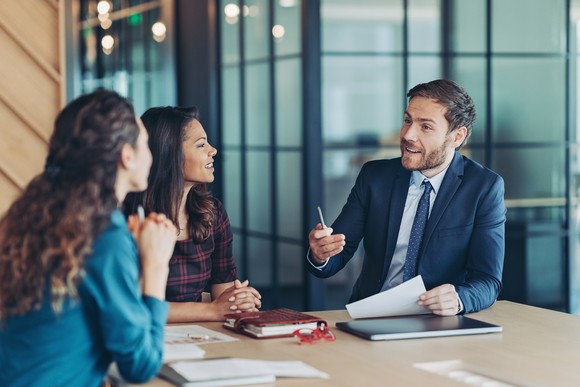 A business person gestures with their hand while holding a piece of paper and engaging with two other people.