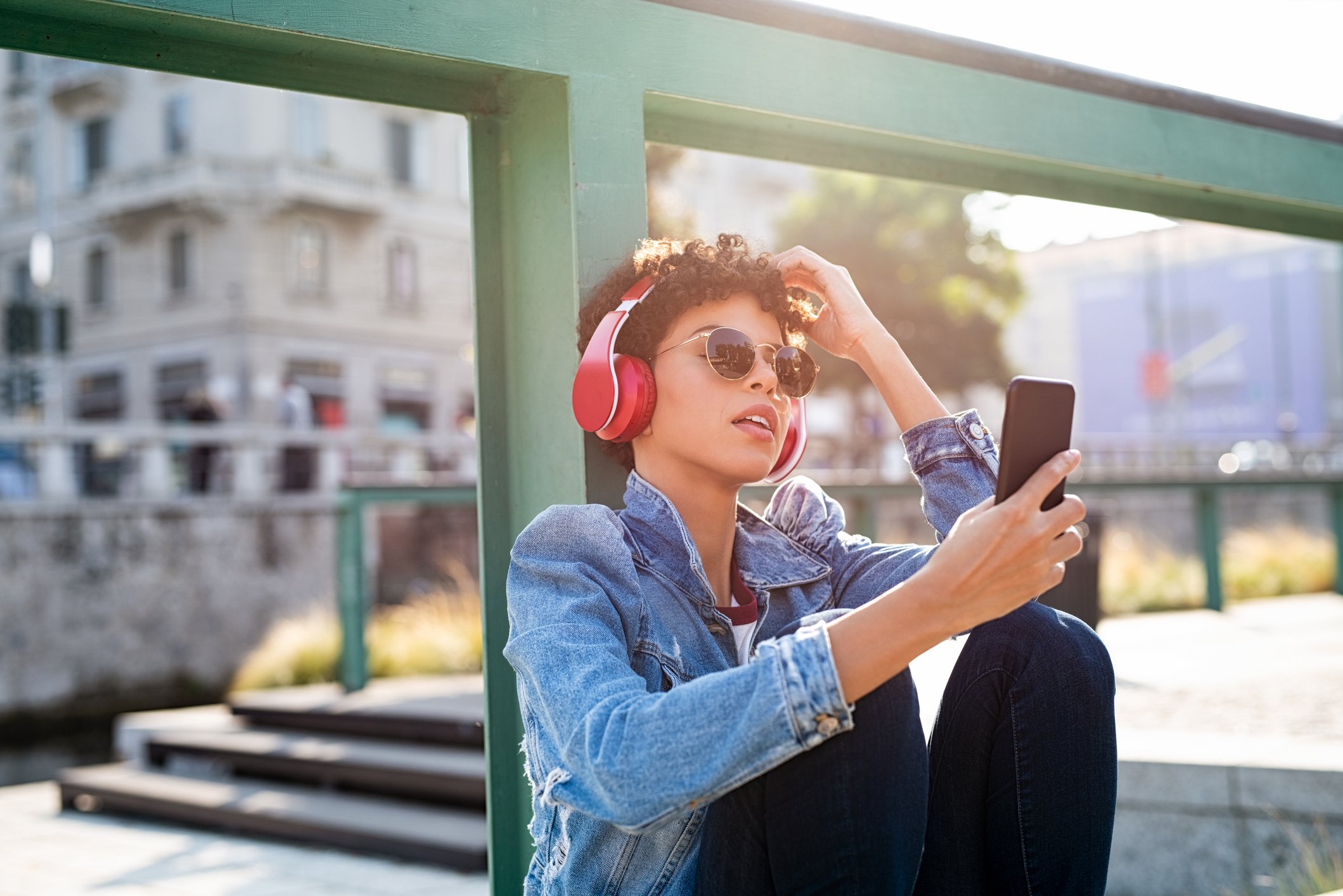A person wearing headphones sitting outdoors looks at a mobile phone.