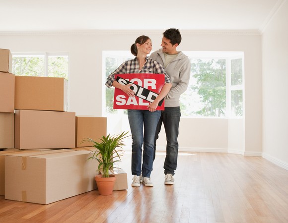 Two people holding for sale sign with the word Sold over it next to boxes in empty living room.