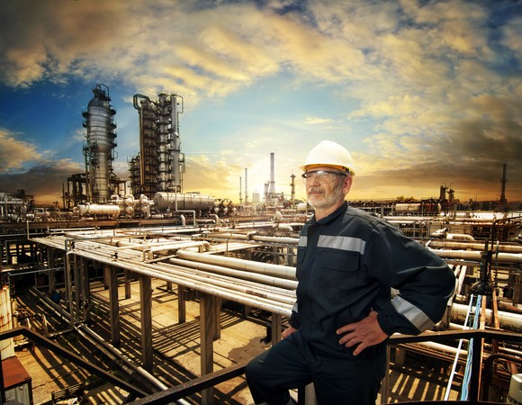 A worker wearing a hard hat with an oil refinery in the background.