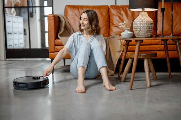 A person sitting on a living room floor activates a robotic vacuum cleaner.
