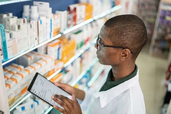 A pharmacist holds a tablet while looking at shelves of medication in a pharmacy.