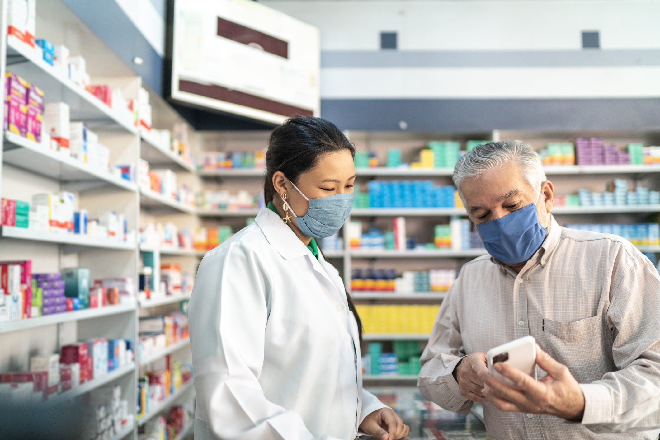 A pharmacist in a pharmacy helps a customer who is showing her some data on his phone.