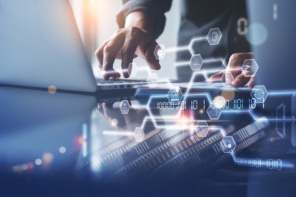 A hand touches a laptop keyboard. In front of it are digital hexagons connected by intersecting lines.