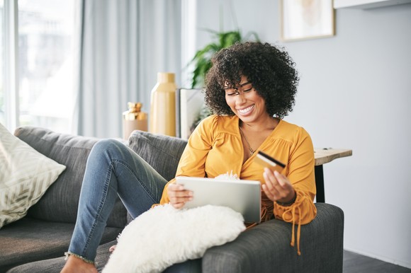 A person smiles and holds a credit card in front of a tablet in a living room setting.