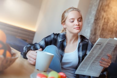 A woman holds a mug while reading the newspaper.