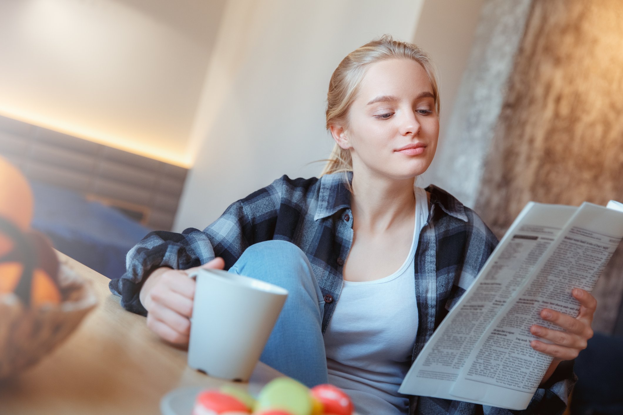 A person holds a newspaper in one hand while sitting at a table and holding a mug.