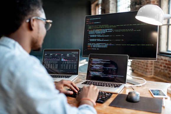 A software engineer writes code on a laptop in front of a larger monitor. 