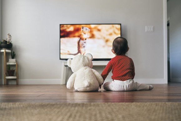 Toddler holding hands with a teddy bear while sitting on the floor and watching TV.
