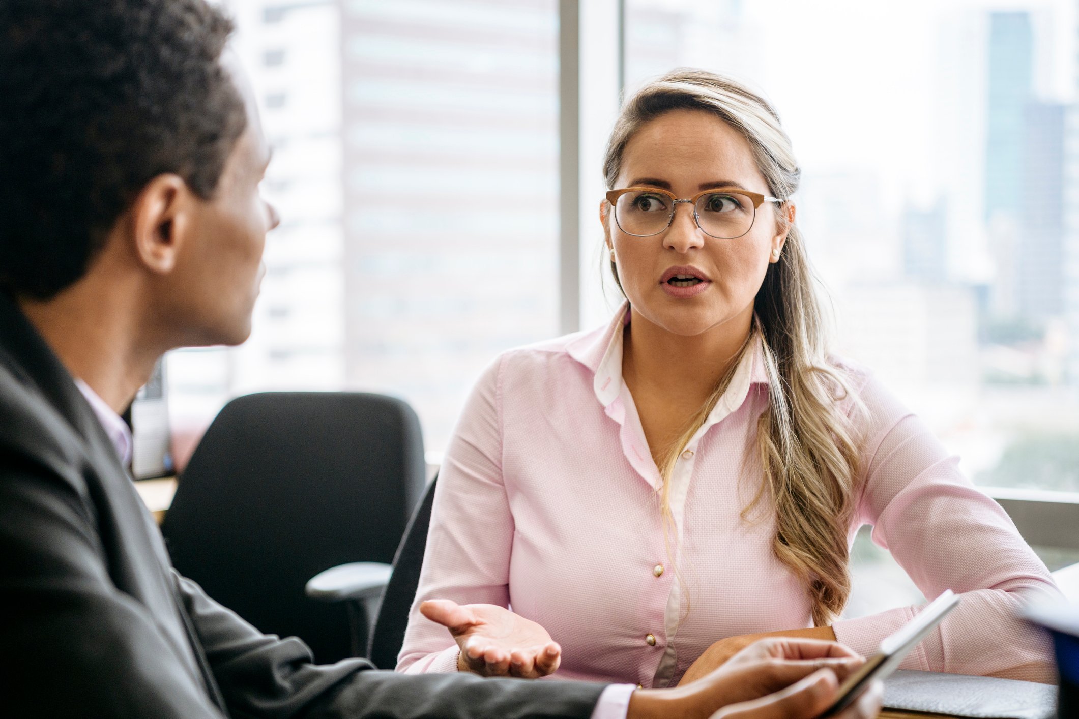 An investor expresses hesitation as she talks to another investor while sitting at a table in an office.