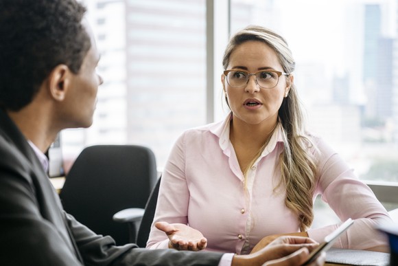 An investor expresses hesitation as she talks to another investor while sitting at a table in an office.