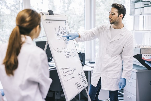 A scientist gestures to a drawing on a whiteboard while in the laboratory with a colleague.