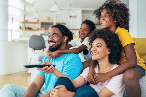 A family sits on a sofa. One person points a remote control.