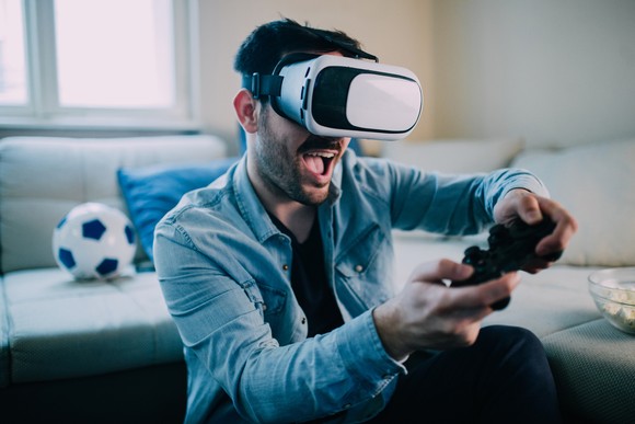 A person wearing a VR headset in a living room plays a game with an excited expression.