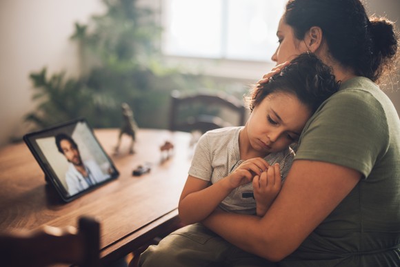 A parent with a child looking at a doctor on a touchscreen tablet.