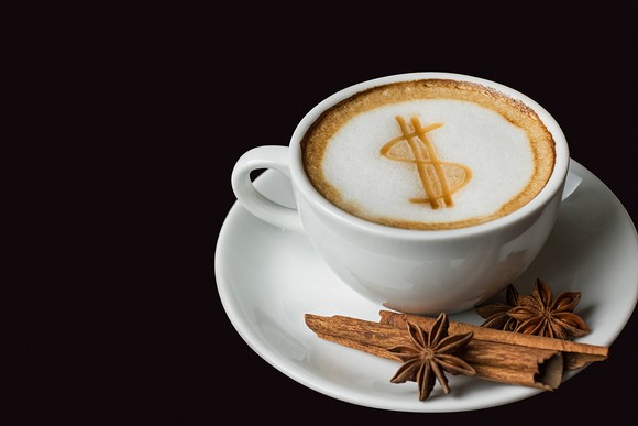 Coffee cup with dollar sign in the foam on the coffee and cinnamon sticks on the saucer. 