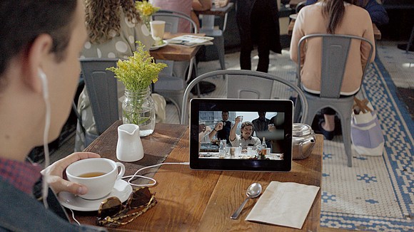 A person sitting at a table holds a coffee cup while watching a streaming service on a tablet computer sitting on the table. The person is wearing earbuds.